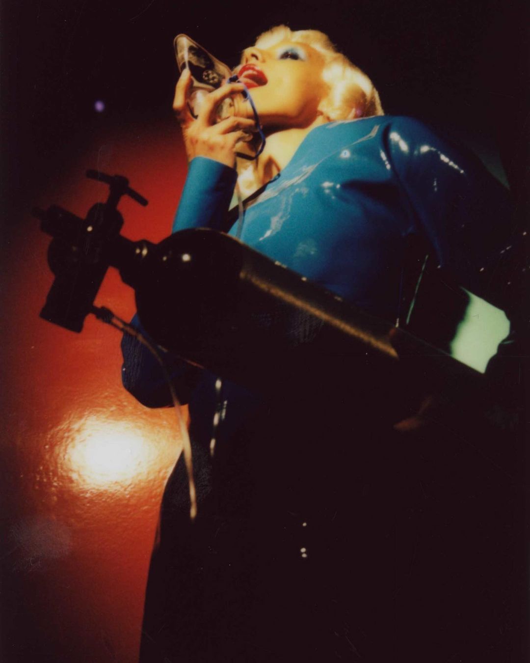 Adult woman in shiny blue outfit poses indoors applying makeup with hand mirror in dark lighting with red highlights, looking up.