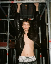 Young man hanging from metal scaffolding wearing an open black leather jacket and printed underwear.