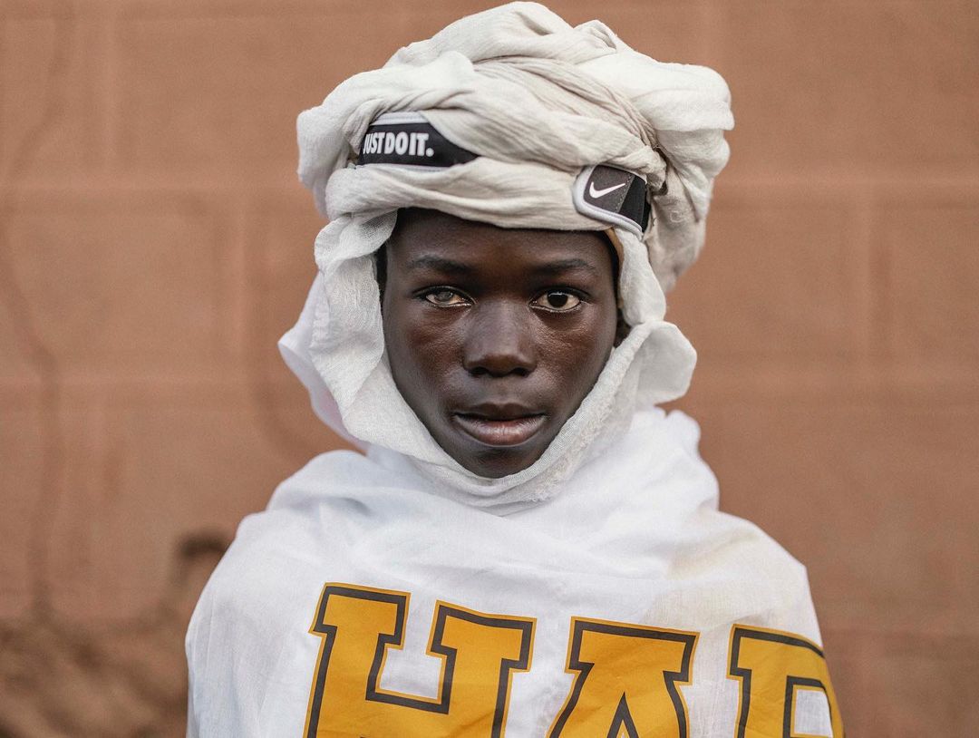 Portrait of a young African man wearing a white turban and white shirt with a yellow graphic, set against a tan brick wall.