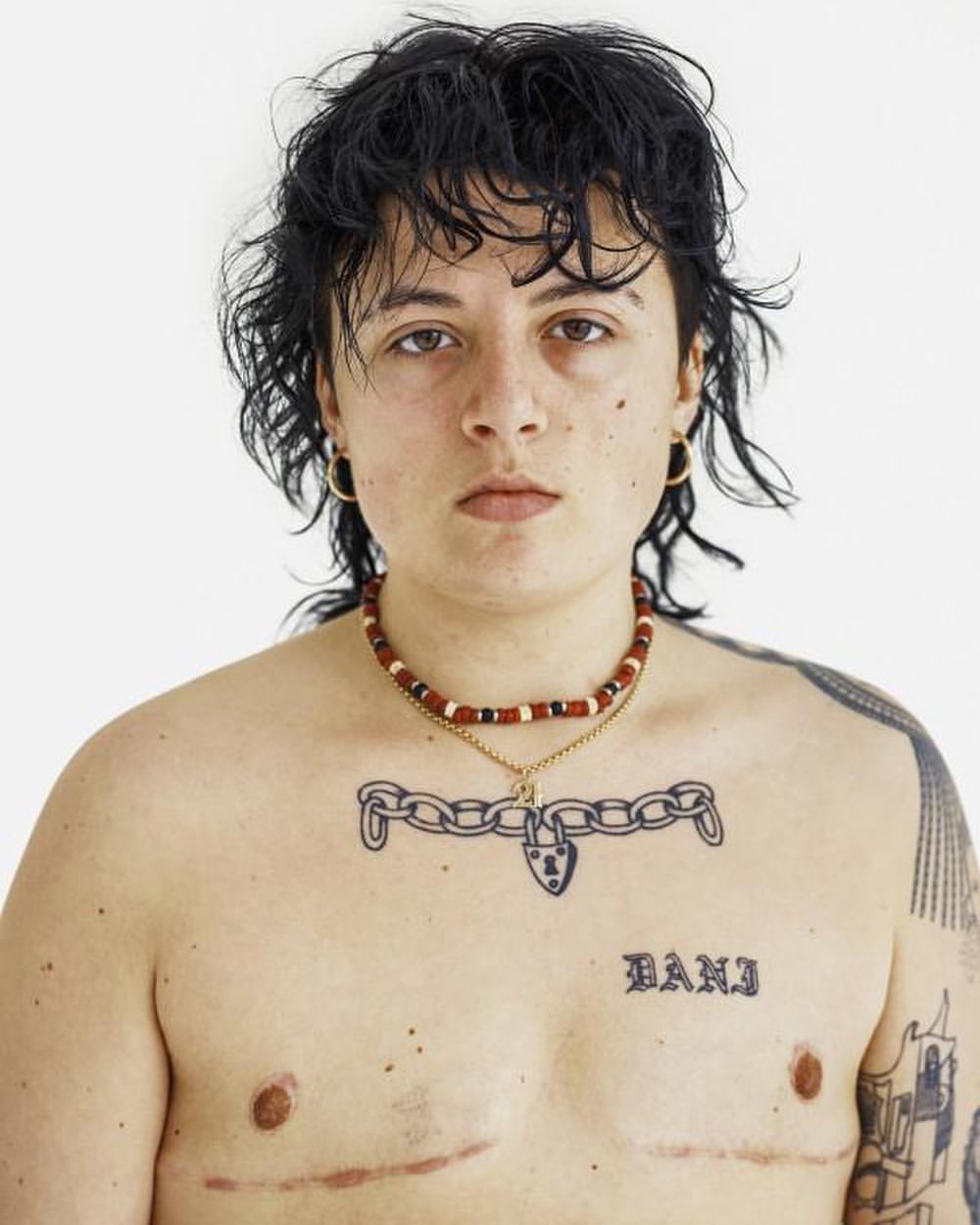 Striking portrait of a shirtless person with a beaded necklace and chest tattoos against a solid white backdrop, studio lighting.