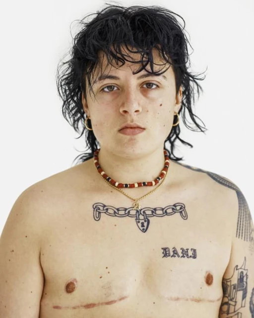 Striking portrait of a shirtless person with a beaded necklace and chest tattoos against a solid white backdrop, studio lighting.