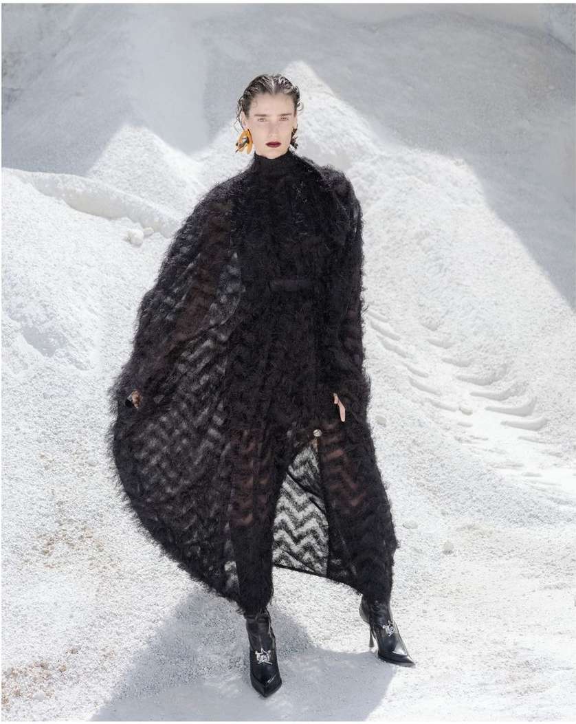 Fashionable woman in a black textured coat poses in a bright, open, snowy environment for a high-fashion editorial photo.