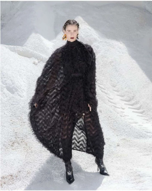 Fashionable woman in a black textured coat poses in a bright, open, snowy environment for a high-fashion editorial photo.
