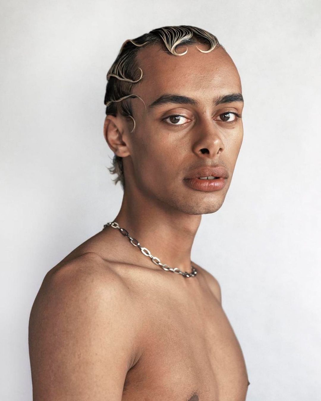 Portrait of a person with dark skin, light eyes, wet-look hair styled in waves, and wearing a silver chain necklace against a white backdrop.