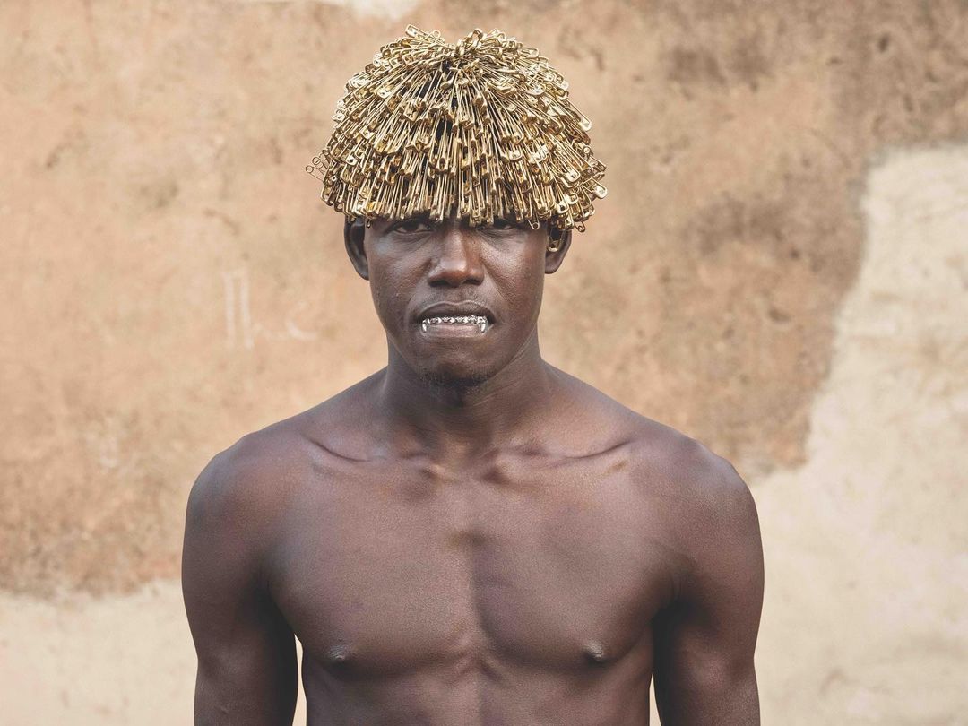 Close-up portrait of a shirtless Black man wearing a hat made of safety pins, set against a neutral textured background.