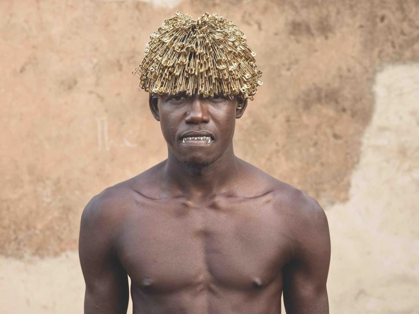 Close-up portrait of a shirtless Black man wearing a hat made of safety pins, set against a neutral textured background.