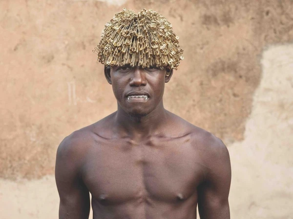 Close-up portrait of a shirtless Black man wearing a hat made of safety pins, set against a neutral textured background.