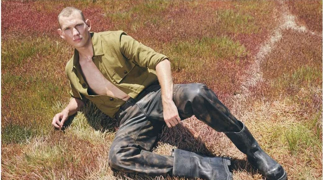 Male fashion model lies in a field wearing an unbuttoned olive drab shirt, black leather pants, and black leather boots in a medium shot.