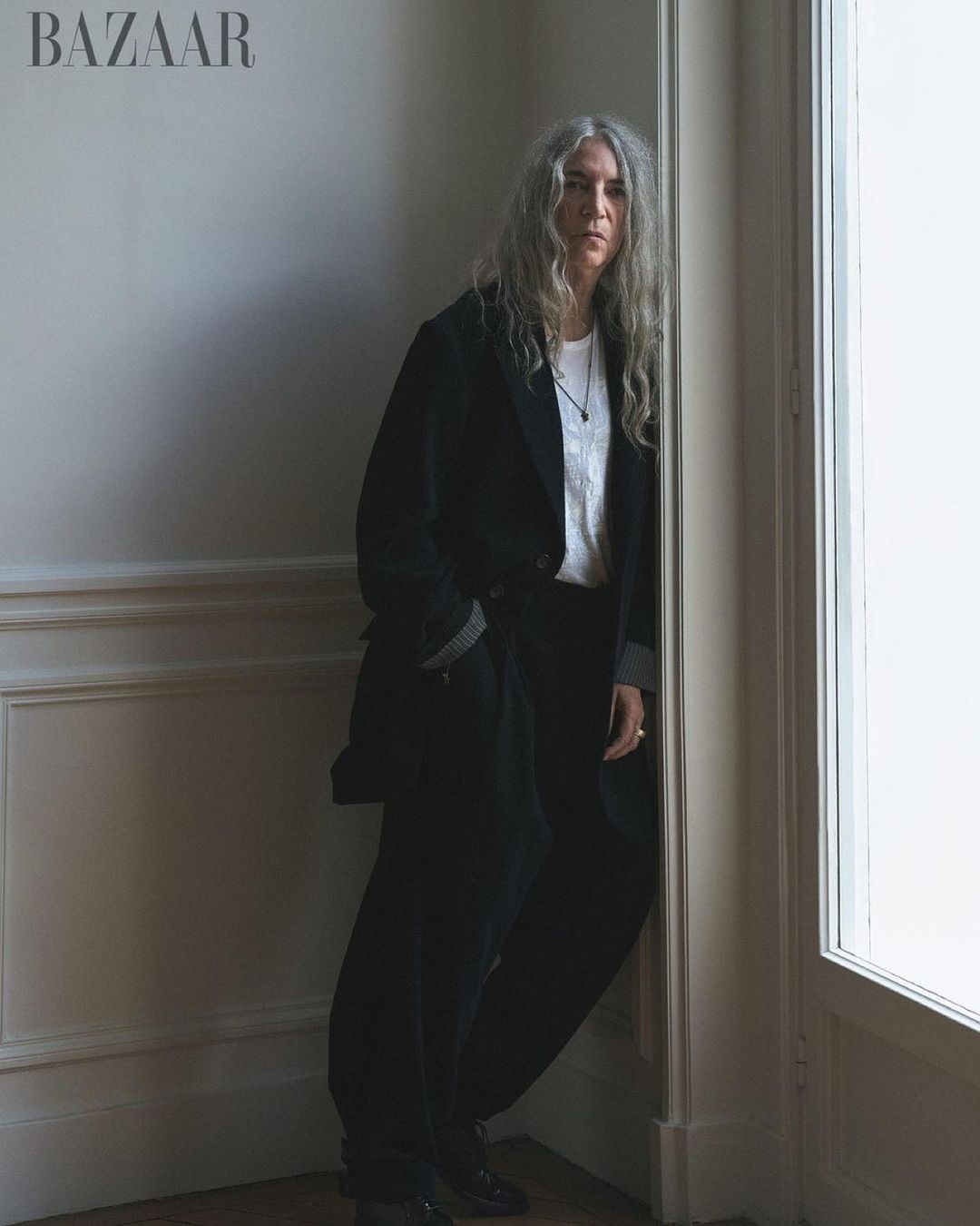 An adult woman with long grey hair leans against a corner in a black blazer, white t-shirt, and black pants in an indoor setting.