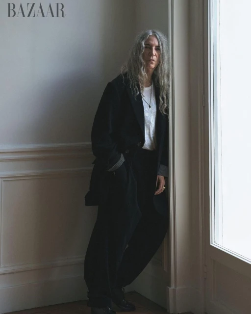 An adult woman with long grey hair leans against a corner in a black blazer, white t-shirt, and black pants in an indoor setting.