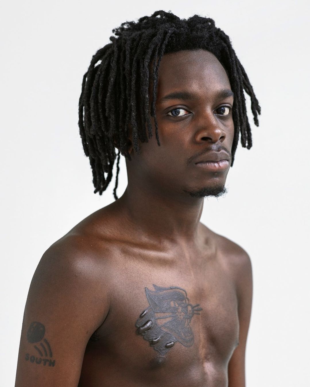Portrait of a young Black man with dreadlocks, tattoo on his chest and arm, against a white background, looking directly at the camera.