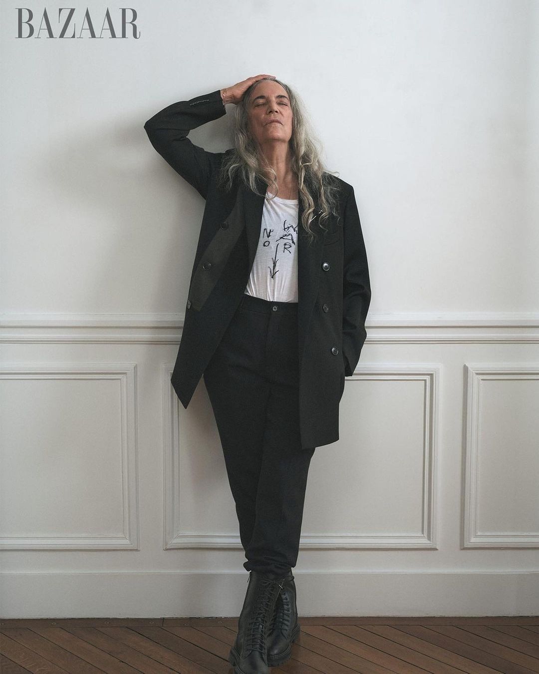 Fashion shot of a mature woman posing in a black pantsuit, with her hand on her head, against a white wall.  Portrait for Bazaar.