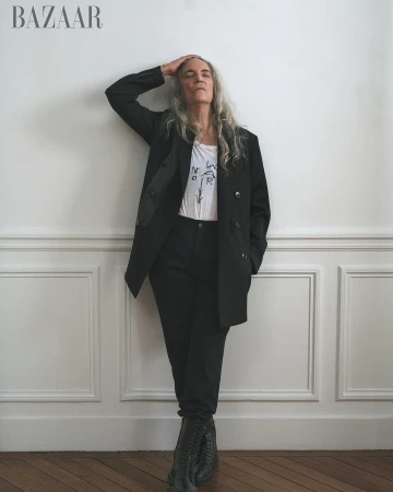 Fashion shot of a mature woman posing in a black pantsuit, with her hand on her head, against a white wall.  Portrait for Bazaar.