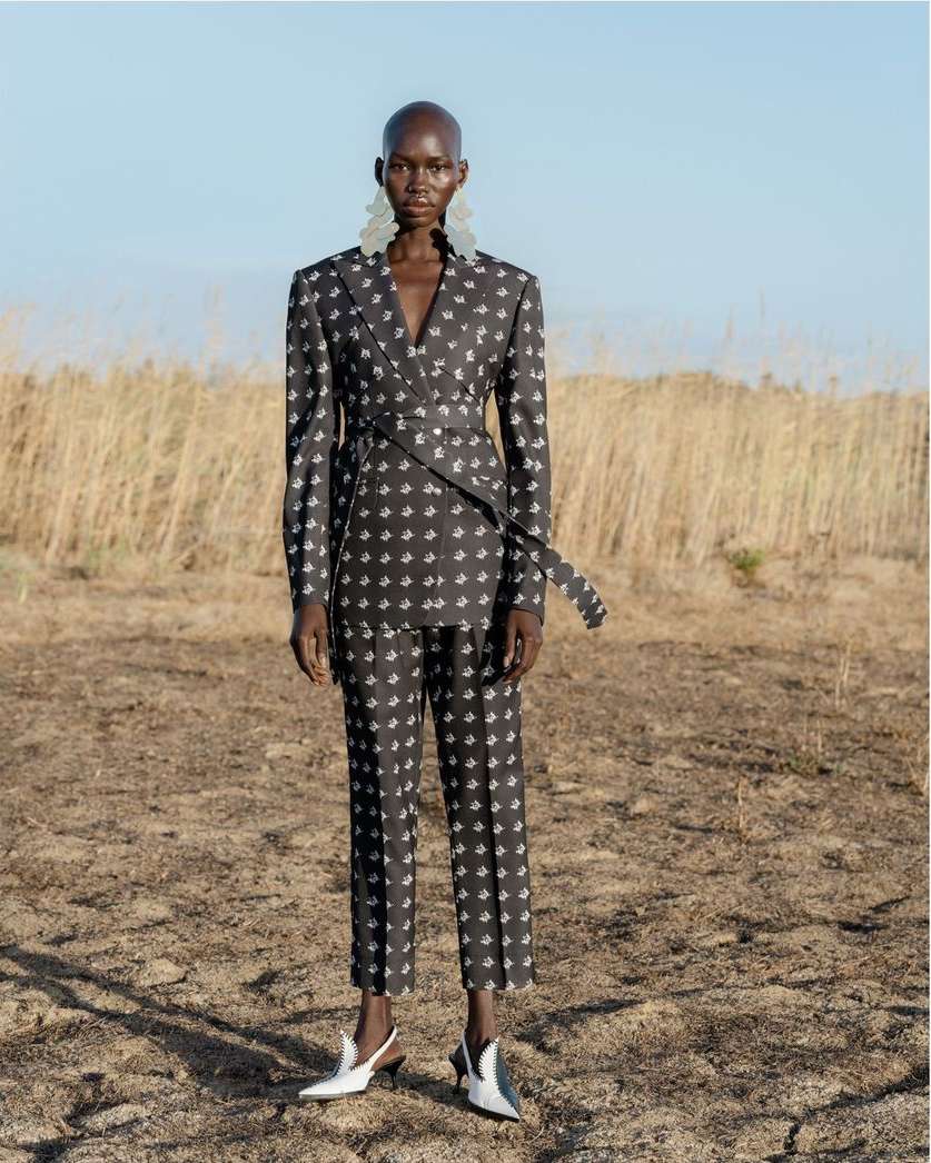 Adult woman poses in patterned suit outdoors with an arid landscape and bright sky behind. She wears statement earrings and pointed flats.