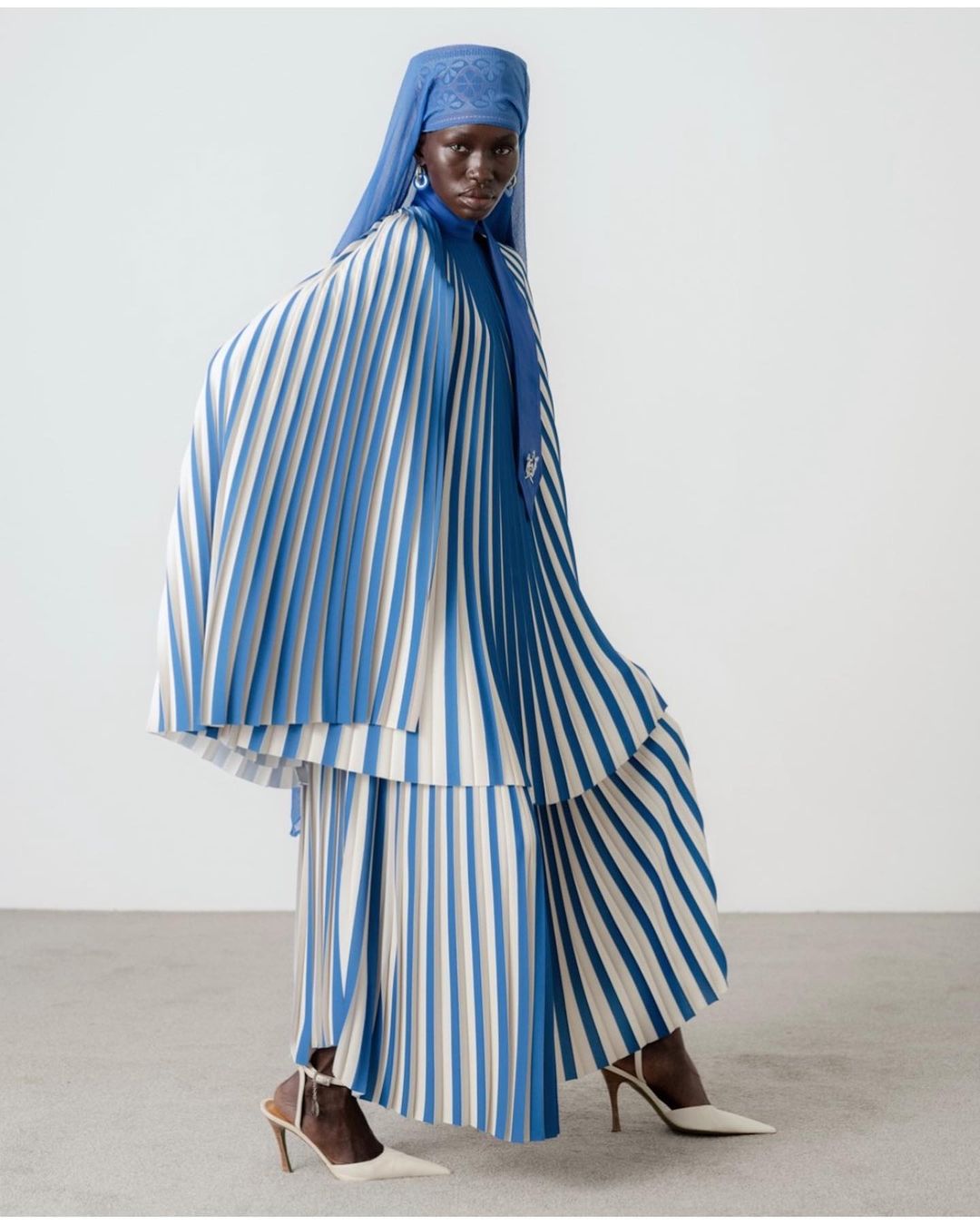 Dark-skinned model posing in a pleated dress with blue and white stripes and a matching headscarf against a white backdrop.