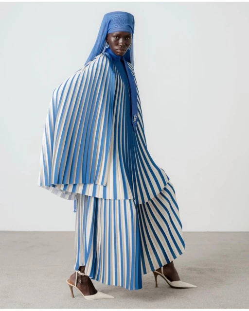 Dark-skinned model posing in a pleated dress with blue and white stripes and a matching headscarf against a white backdrop.