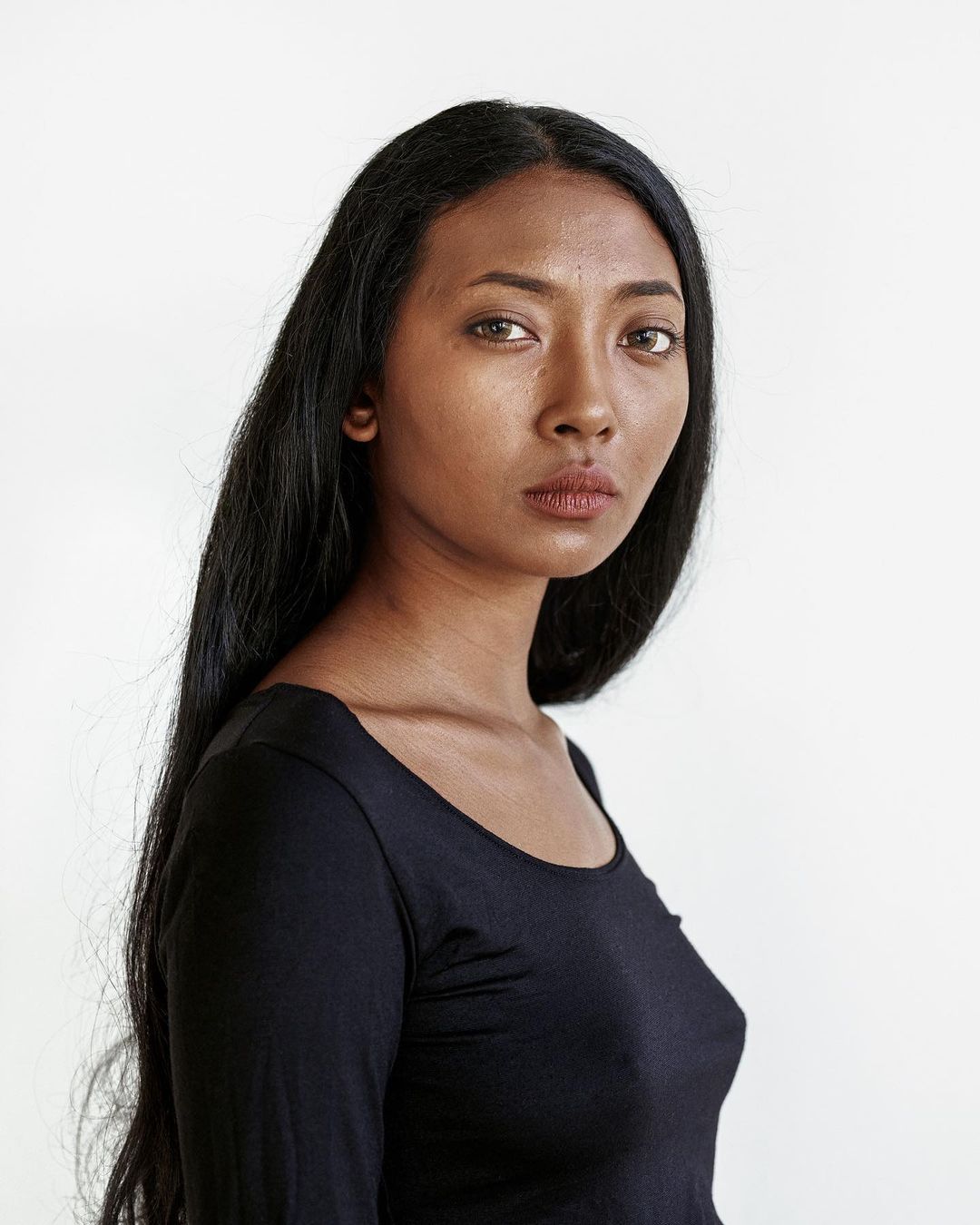 Portrait of a woman with long black hair in a dark top against a white backdrop, looking directly at the camera in a photo studio.