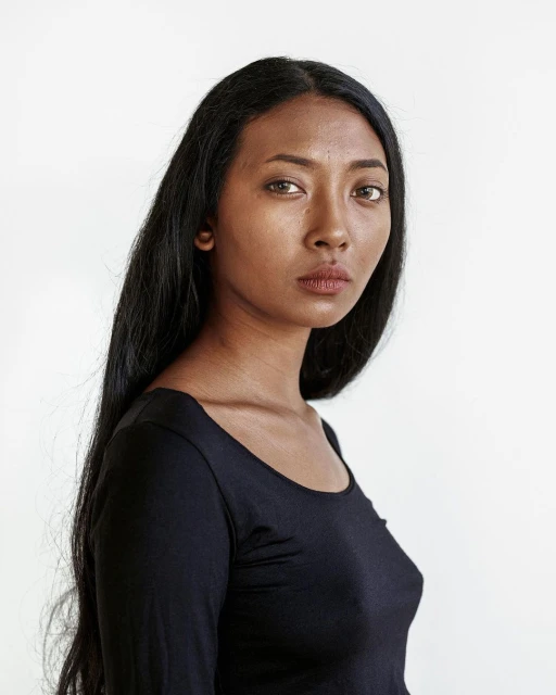 Portrait of a woman with long black hair in a dark top against a white backdrop, looking directly at the camera in a photo studio.