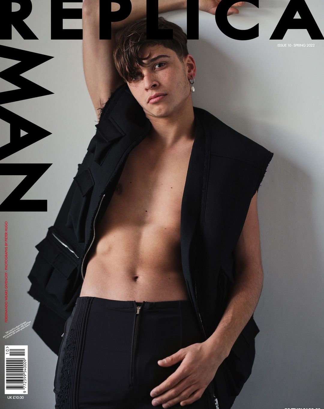 Young man in black sleeveless vest and dark trousers posing against an off-white wall for a magazine cover.