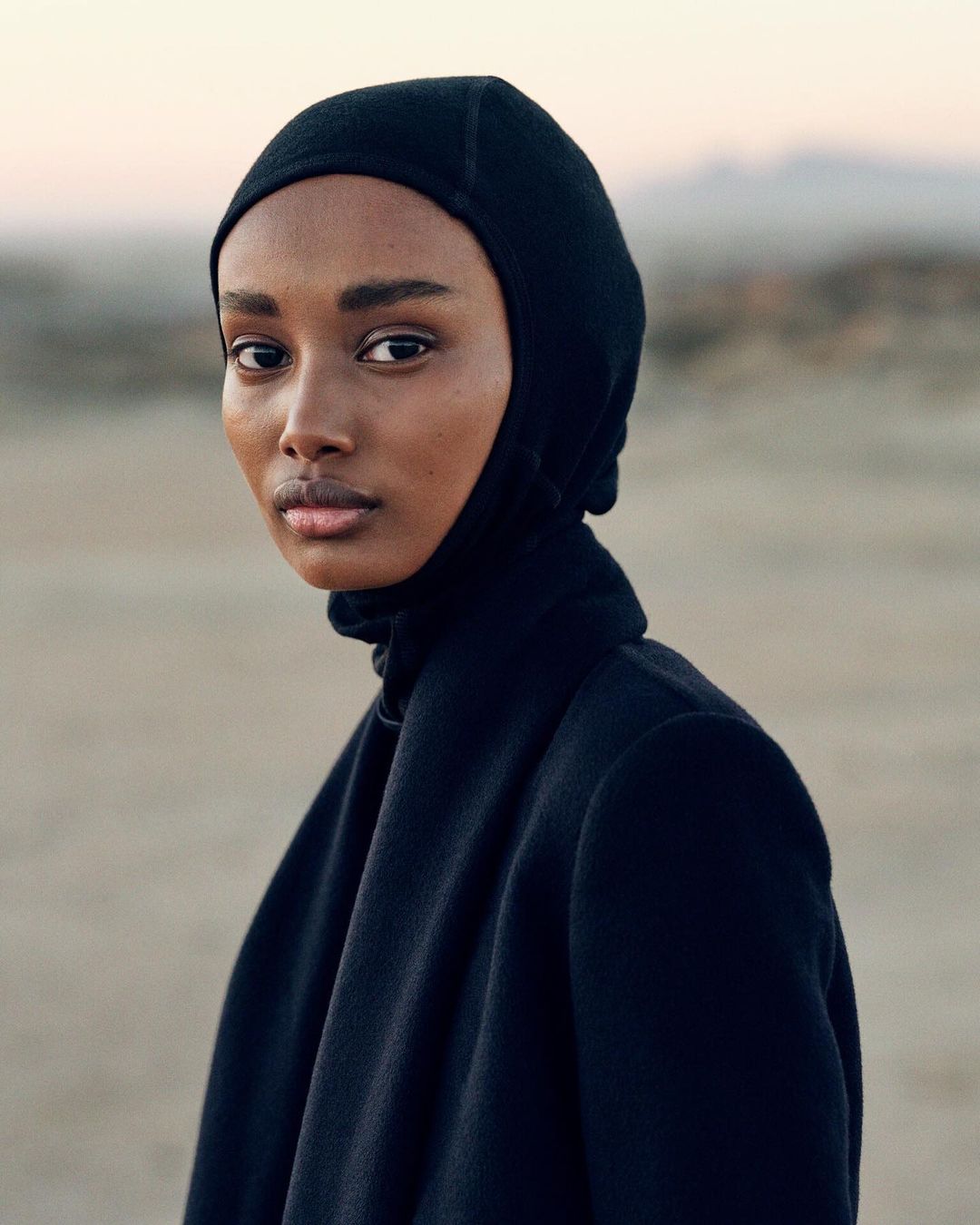 Portrait of a Black female adult with neutral expression, wearing a black balaclava and jacket against a neutral outdoor background.