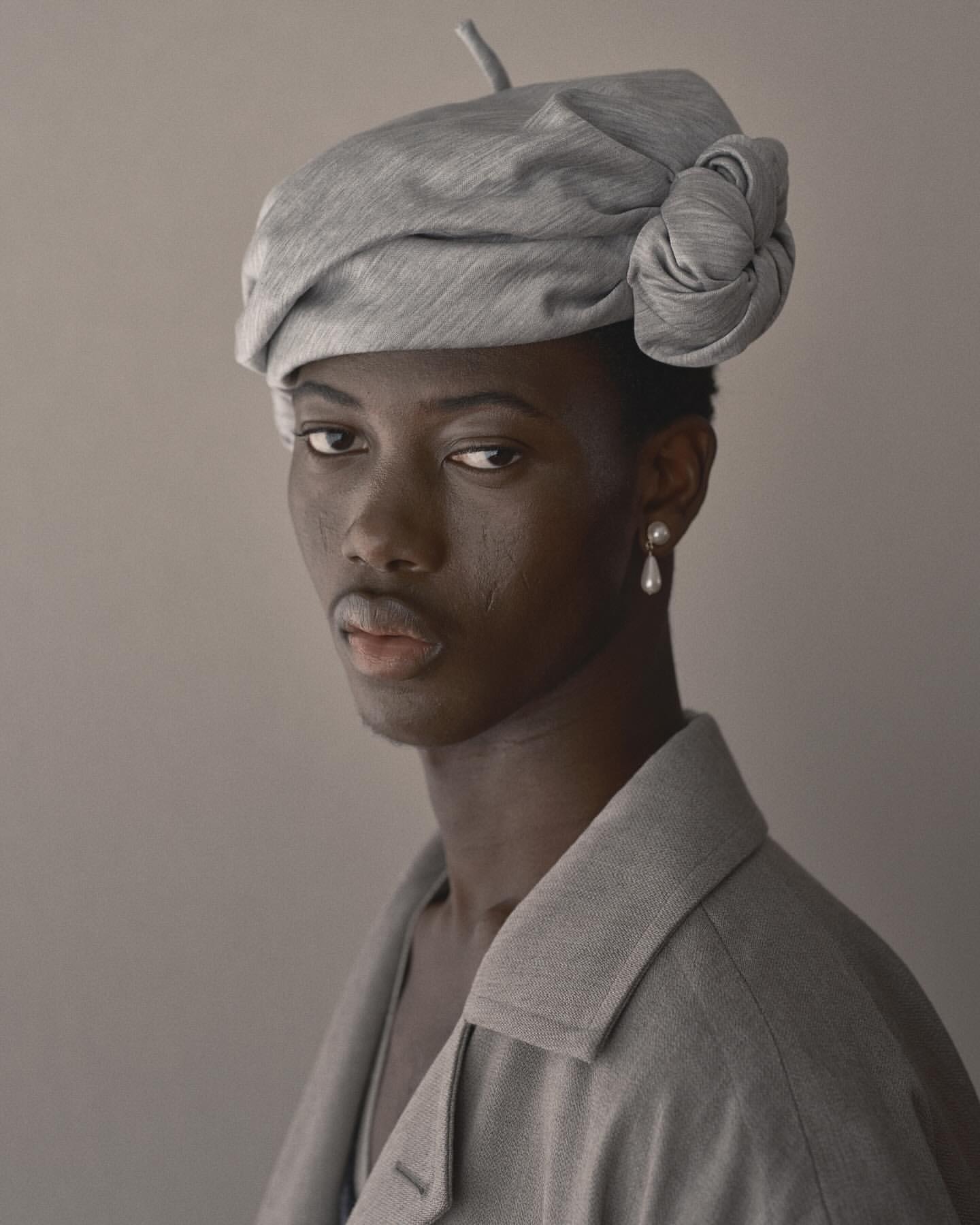 Close-up studio portrait of a Black adult female model wearing a stylish gray beret and matching blazer against a neutral gray backdrop.