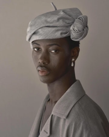 Close-up studio portrait of a Black adult female model wearing a stylish gray beret and matching blazer against a neutral gray backdrop.