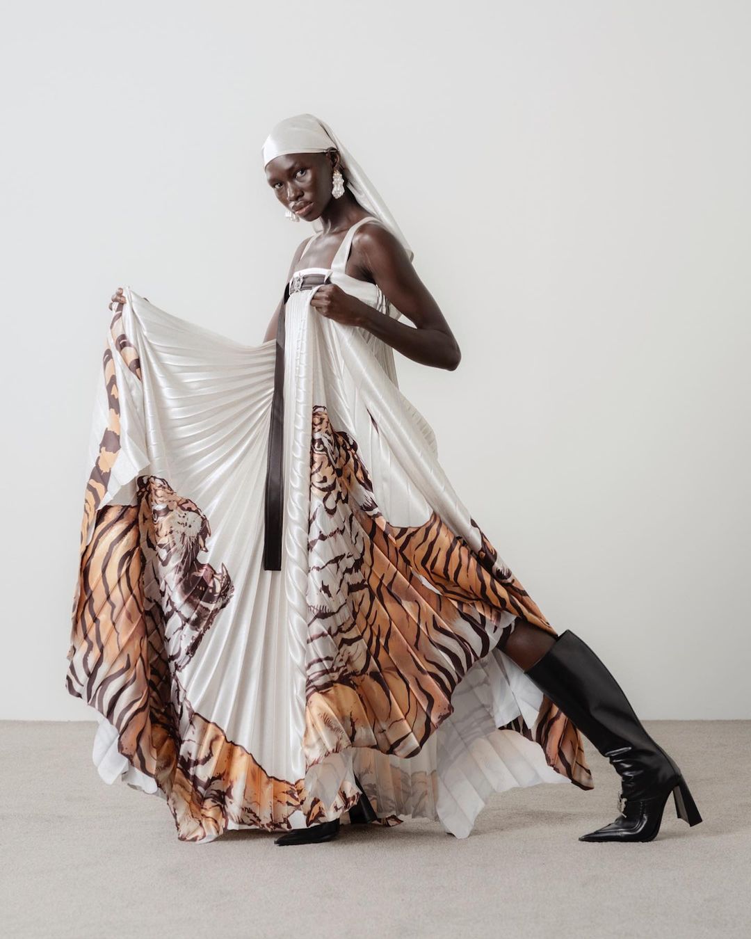Fashion shot of Black woman in white pleated gown with tiger graphics, headscarf, earrings, and boots posing in a photo studio.
