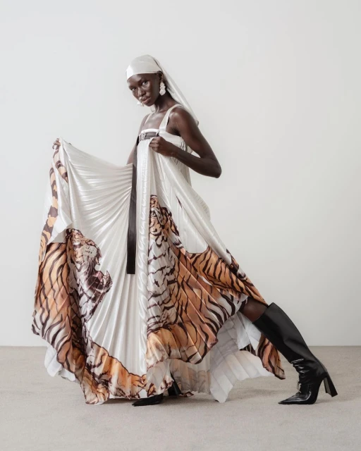 Fashion shot of Black woman in white pleated gown with tiger graphics, headscarf, earrings, and boots posing in a photo studio.