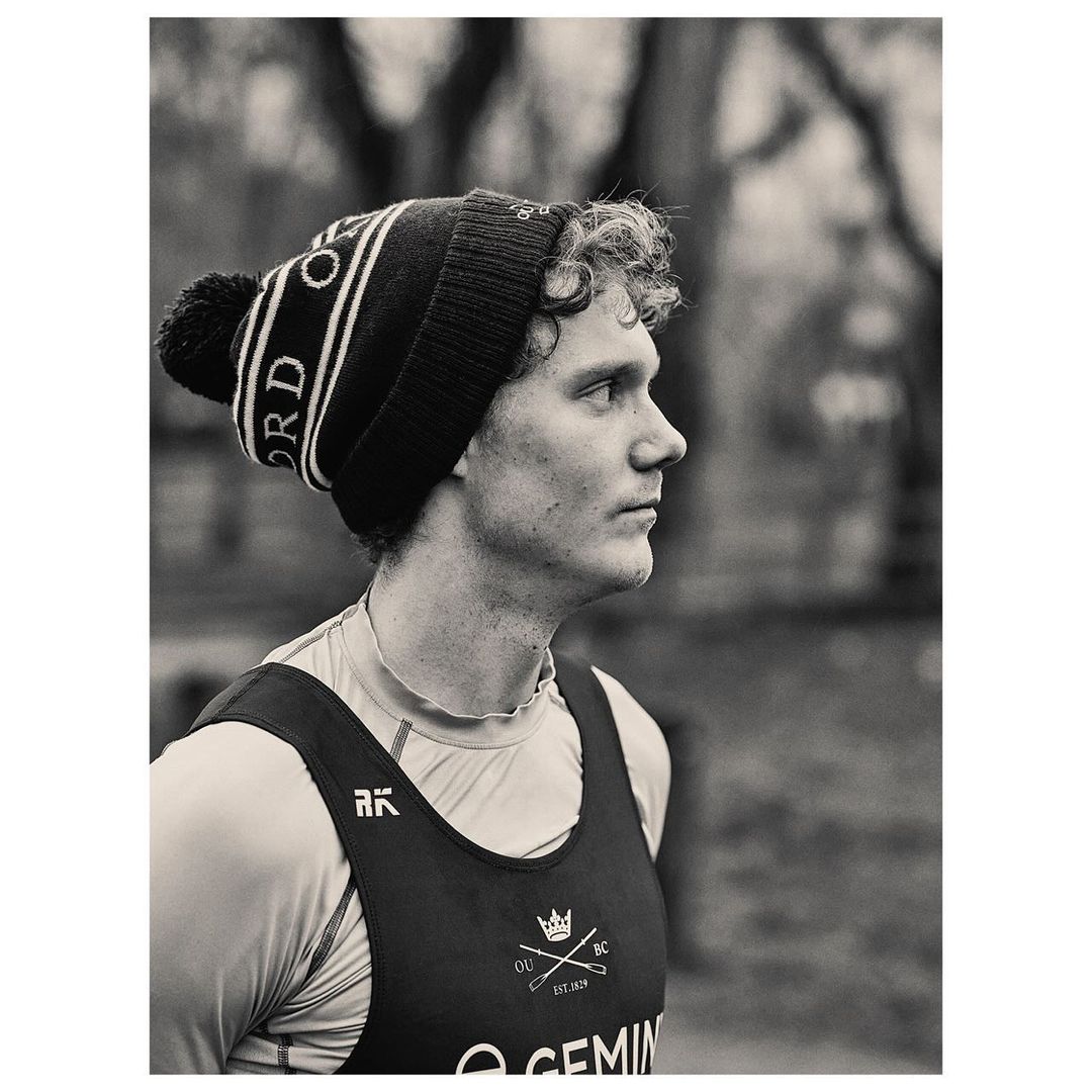 Black and white portrait of an adult male rower with curly hair wearing a branded beanie and rowing uniform outside on a cloudy day.