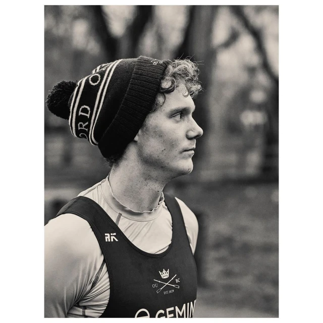 Black and white portrait of an adult male rower with curly hair wearing a branded beanie and rowing uniform outside on a cloudy day.