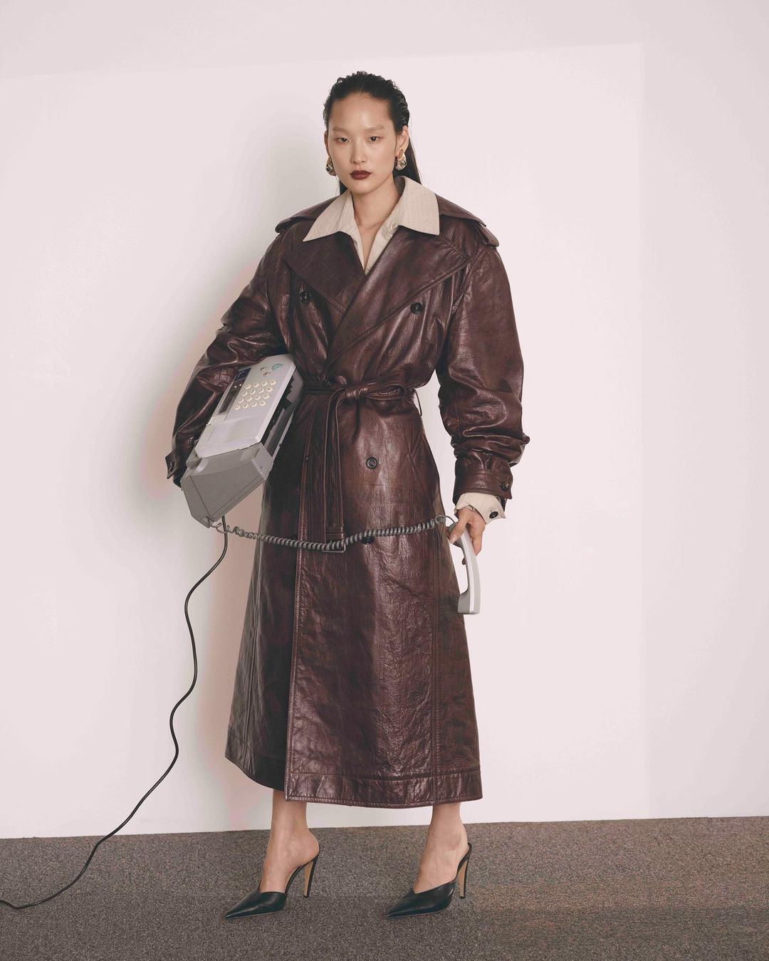 A full-length portrait of an Asian woman in a brown leather trench coat holding a retro telephone in a brightly lit indoor studio.