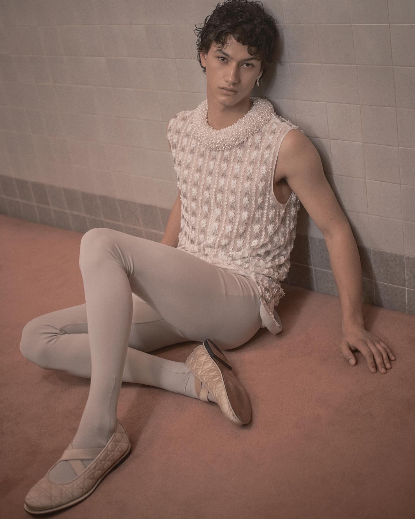 A young adult male in a light sleeveless top and leggings, seated on a pink floor, leaning against a tiled wall in a minimalist style.