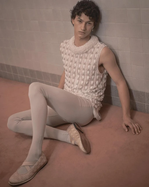 A young adult male in a light sleeveless top and leggings, seated on a pink floor, leaning against a tiled wall in a minimalist style.