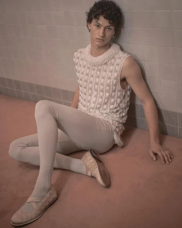 A young adult male in a light sleeveless top and leggings, seated on a pink floor, leaning against a tiled wall in a minimalist style.