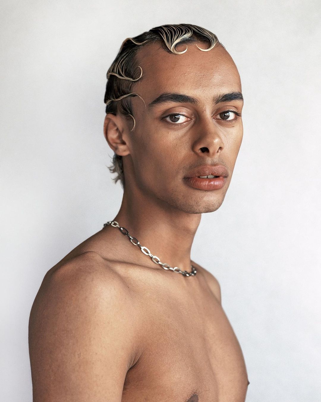 A shirtless person with a silver necklace, brown skin, and wavy blonde hair poses in a studio with a white background, portrait-style.