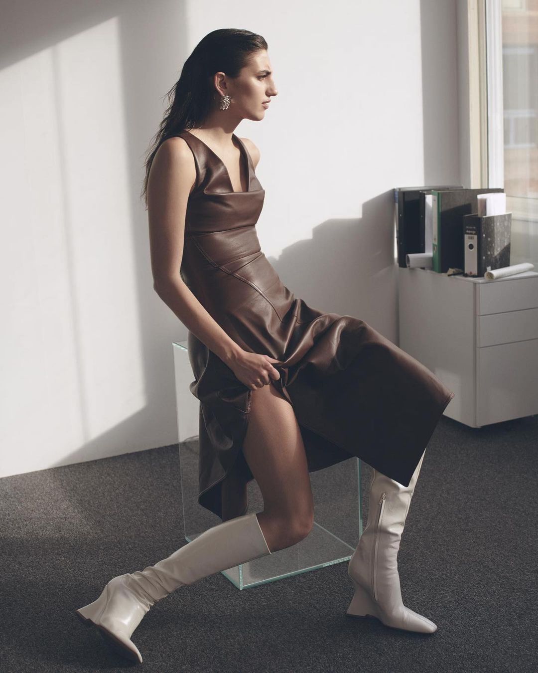 A woman in a brown leather dress sits on a clear cube in a studio, wearing white boots and looking off to the side with neutral expression.