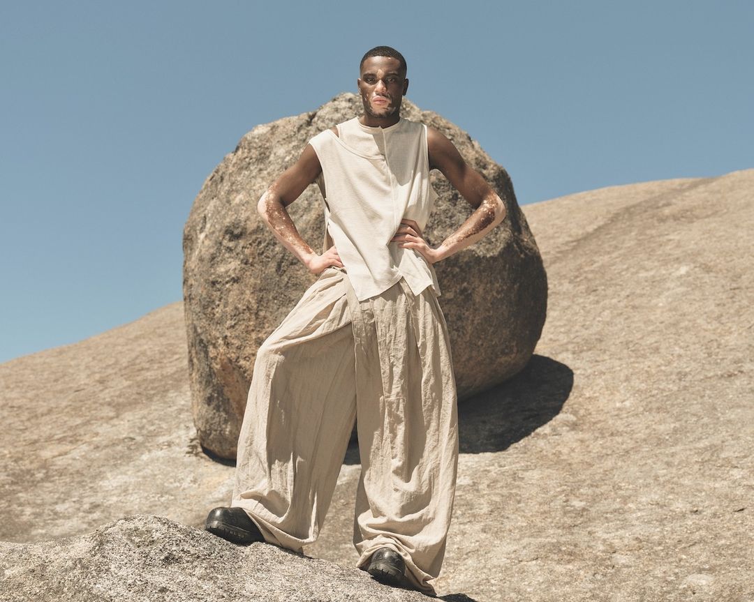 An adult male model poses in tan clothing on a rocky desert landscape with a boulder, under a clear blue sky, for a fashion editorial.