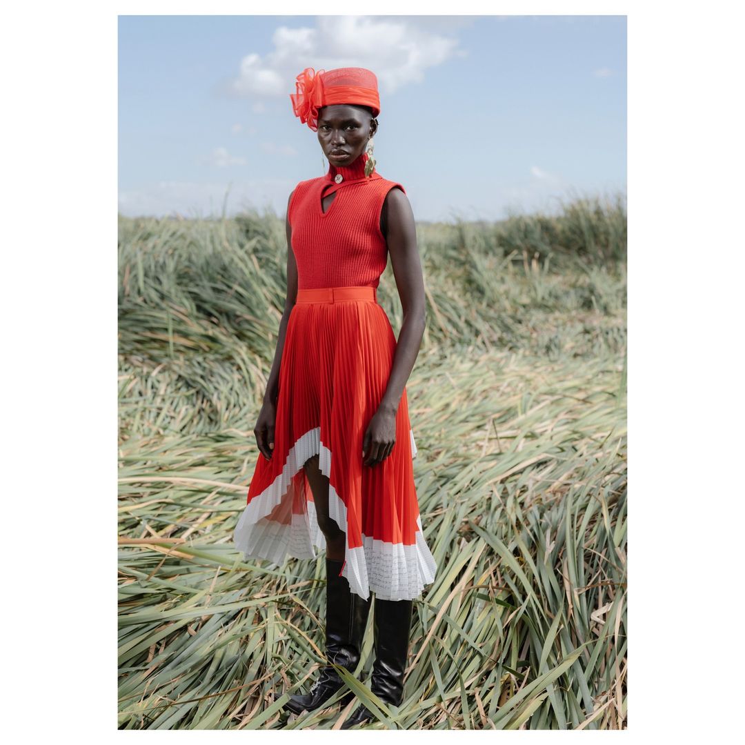 A South Sudanese woman models red high-low pleated dress and matching hat in a grassy field under a blue sky with scattered clouds.