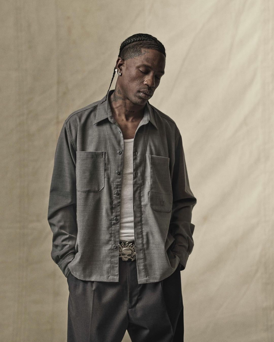 Portrait of African-American man with braids wearing a grey work shirt and black pants, posing against a tan backdrop in a studio.