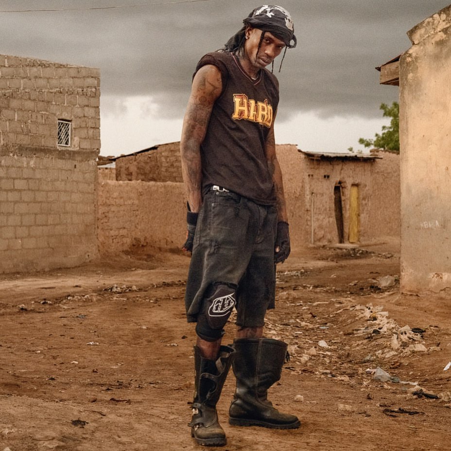 Adult male in work boots and motocross gloves poses in an urban alleyway next to dilapidated buildings on a cloudy overcast day.