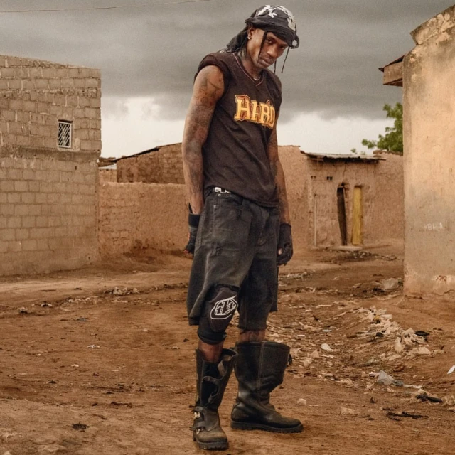 Adult male in work boots and motocross gloves poses in an urban alleyway next to dilapidated buildings on a cloudy overcast day.
