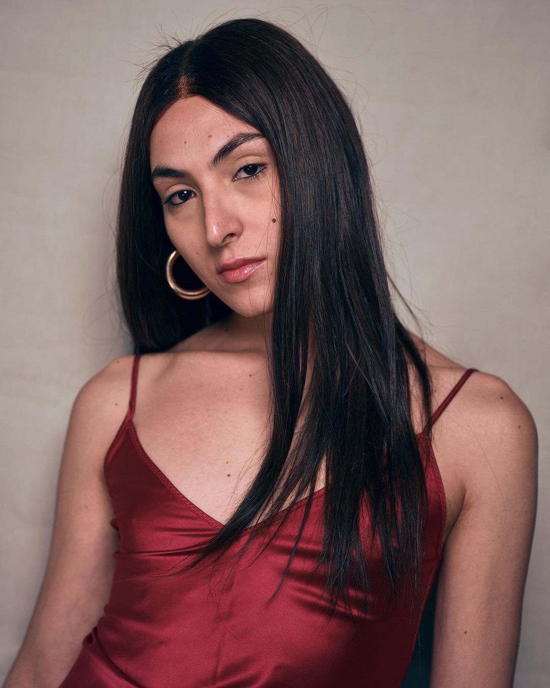 Portrait of a woman with long black hair and gold hoop earrings, wearing a burgundy satin dress against a neutral background.