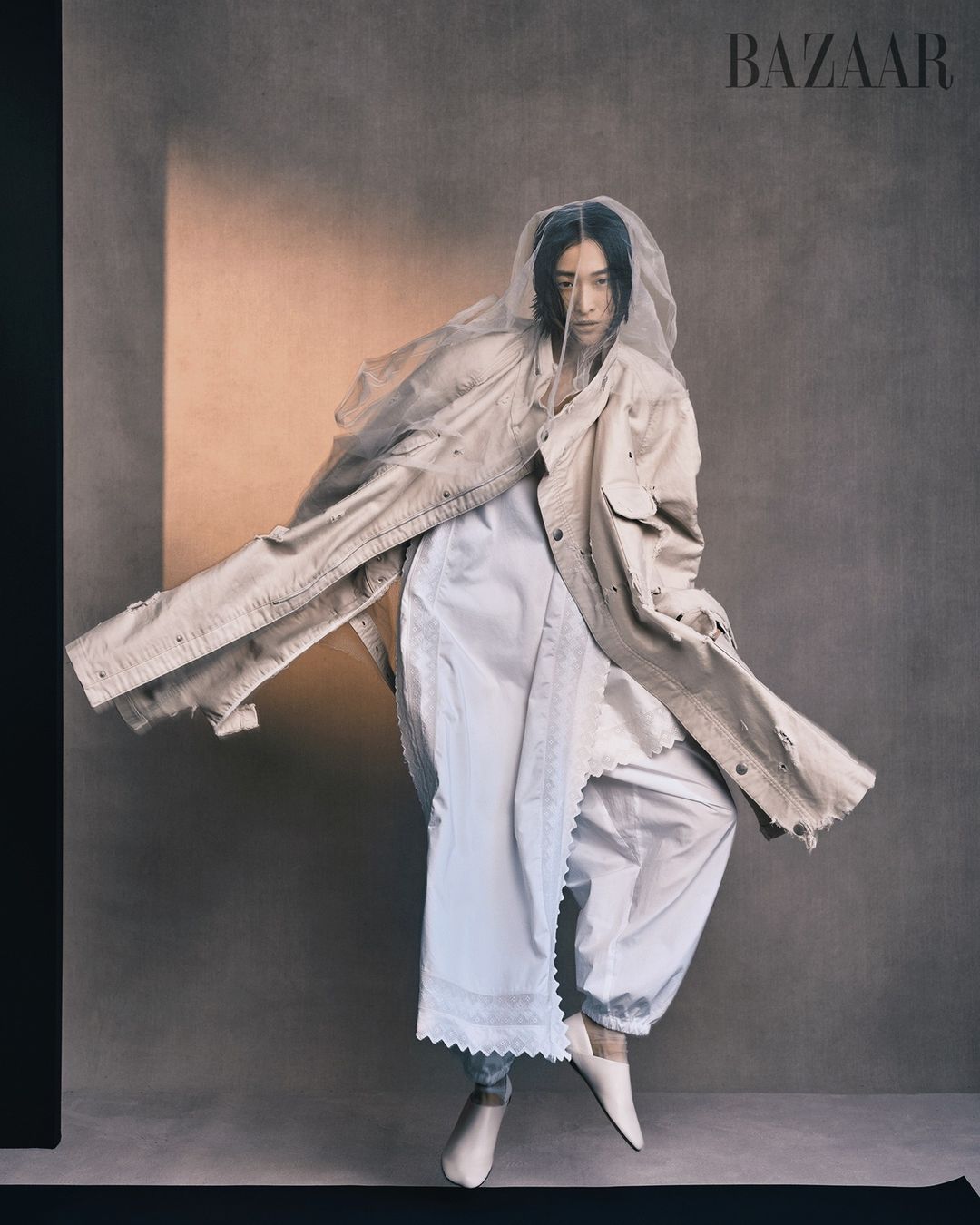 Fashion editorial shot of androgynous model in oversized beige jacket, white skirt, sheer veil, and white heels against grey background.