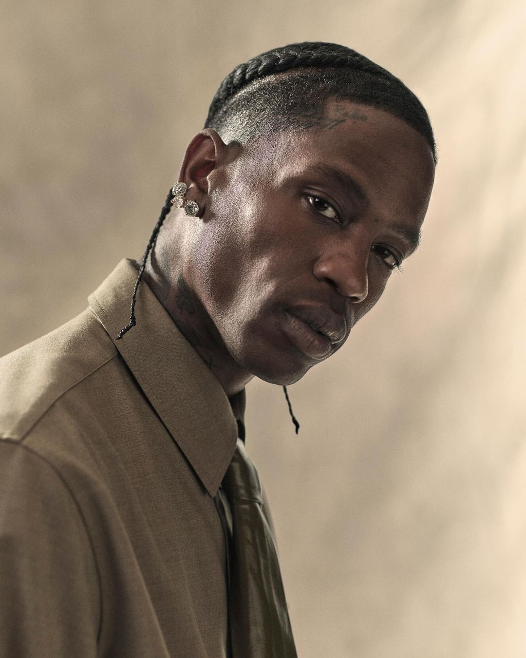 Portrait of an adult man with cornrow braids, wearing a khaki button-down shirt and tie, against a neutral studio backdrop with soft lighting.