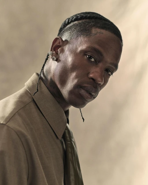 Portrait of an adult man with cornrow braids, wearing a khaki button-down shirt and tie, against a neutral studio backdrop with soft lighting.