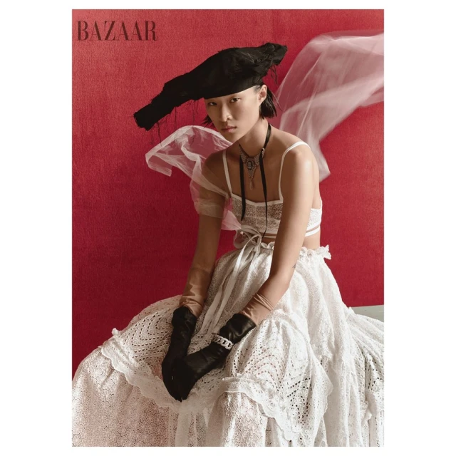 Fashion editorial shot of an Asian woman in an eye-level portrait wearing a white lace dress, gloves and a hat, posed against a red backdrop.