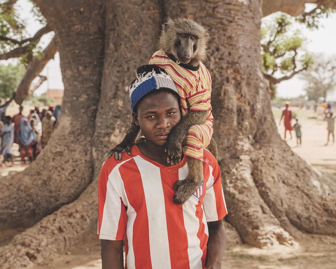 A young man in a soccer jersey carries a baboon on his shoulders in a rural outdoor setting with a baobab tree in the background.