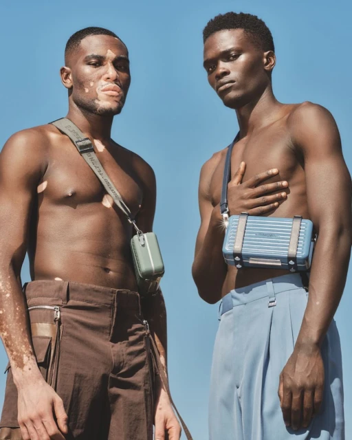 Two bare-chested adult male fashion models pose outdoors against a blue sky, each with a small luggage bag worn as a crossbody purse.