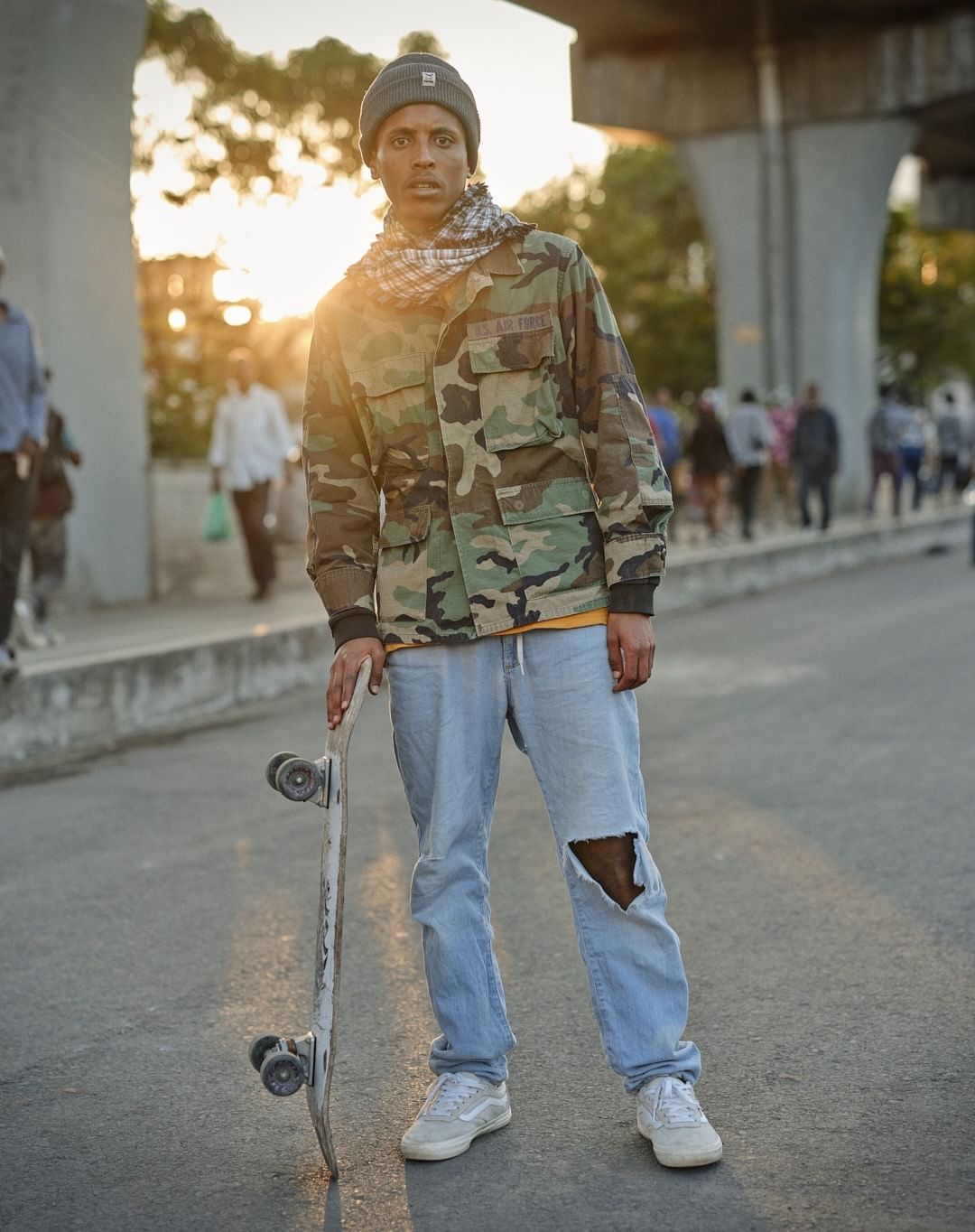 Adult male in camouflage jacket stands on city street with a skateboard in the sunset. He wears a beanie and ripped jeans.