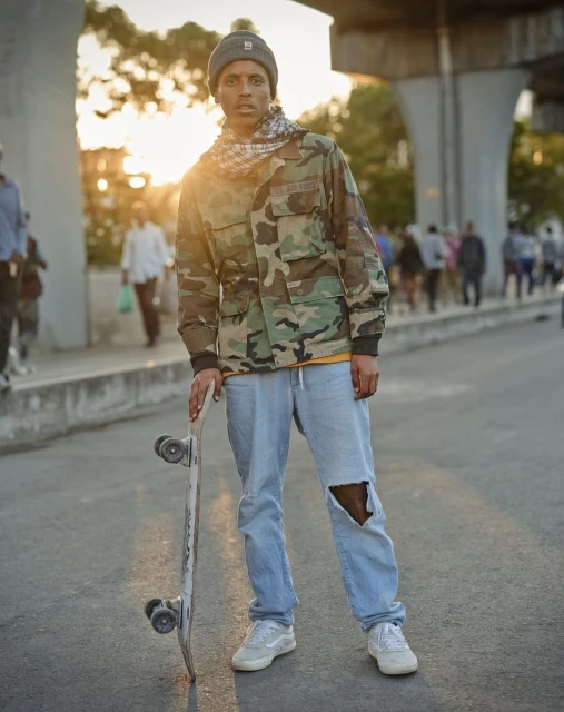 Adult male in camouflage jacket stands on city street with a skateboard in the sunset. He wears a beanie and ripped jeans.